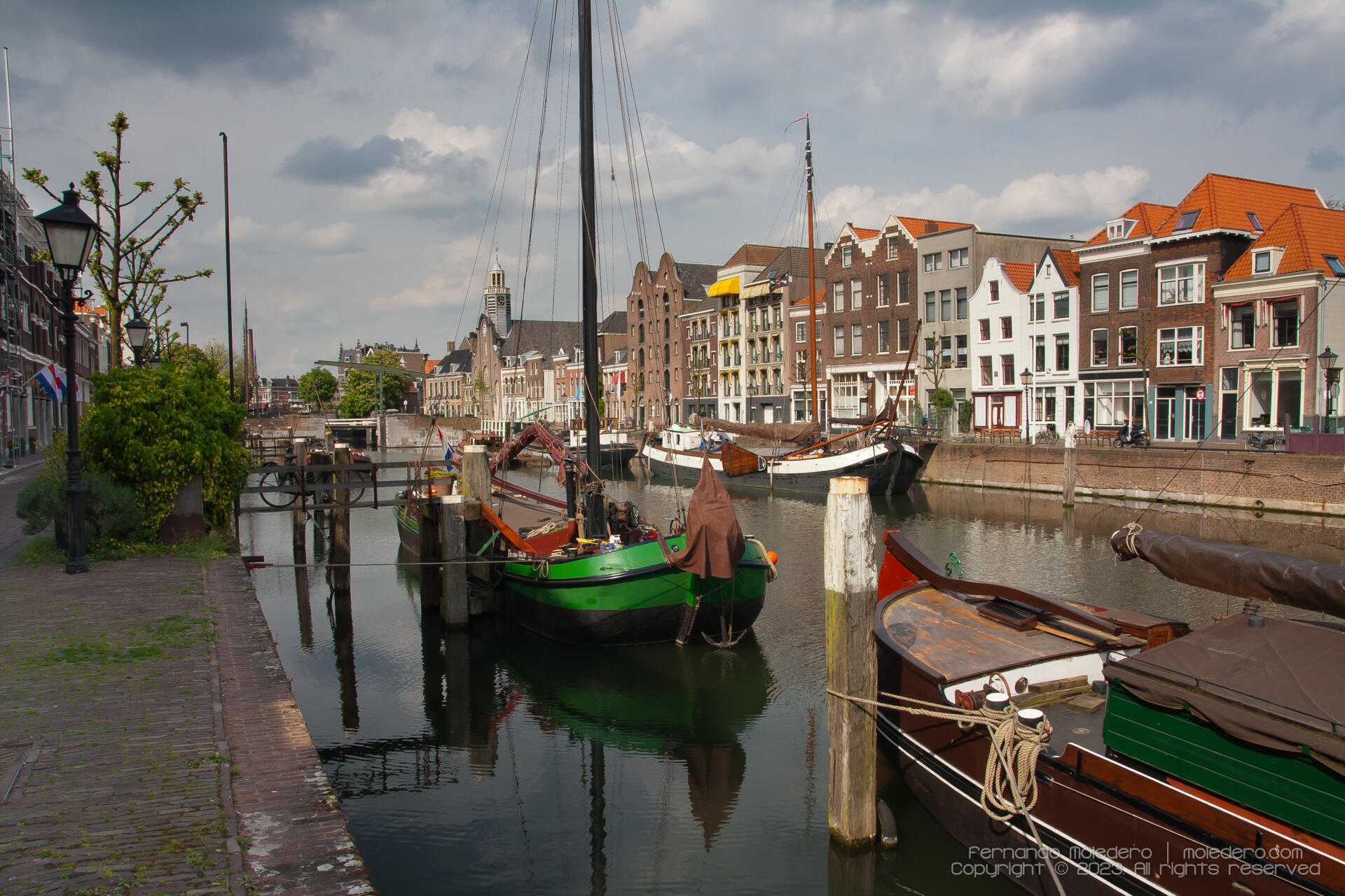 View of Delfshaven harbor in Rotterdam, the Netherlands, showing historic boats, brick gabled houses, and the canal under a partly cloudy sky.