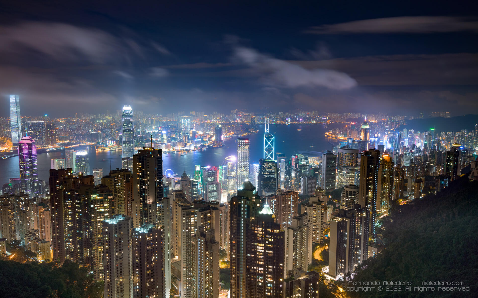 Panoramic night view of Hong Kong, from Victoria Peak, showing the illuminated skyline with skyscrapers, Victoria Harbour and colorful city lights under a cloudy sky