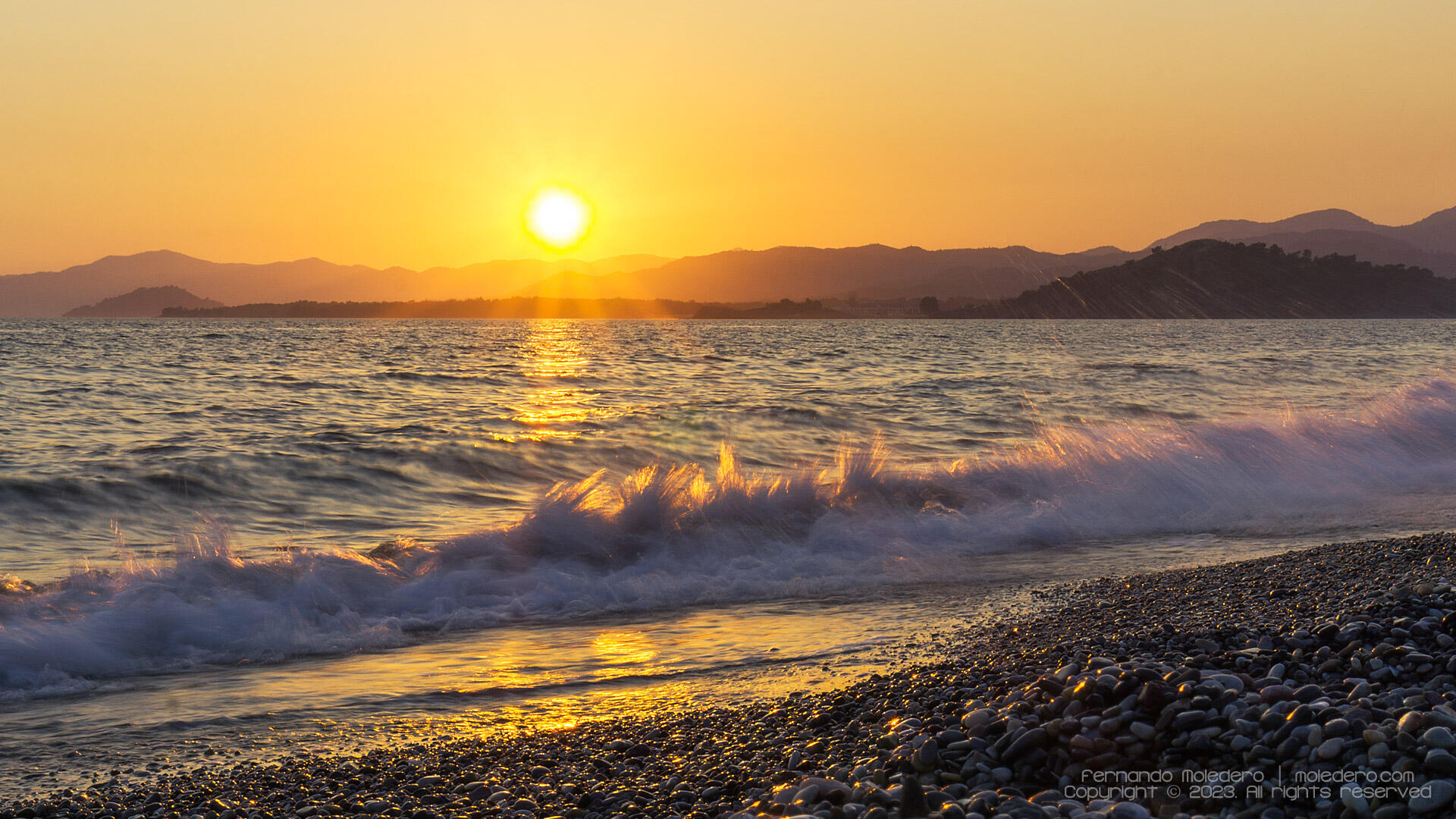 Sunset over the sea at Calis Beach in Fethiye, Turkey, with waves breaking on the pebbled shore