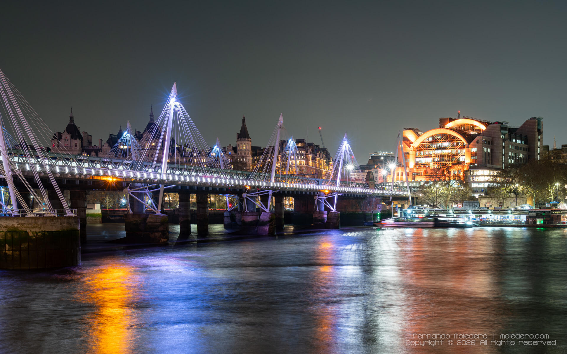 Night view of Hungerford Bridge and the Golden Jubilee Bridges crossing the River Thames, with the illuminated façade of Charing Cross Station behind