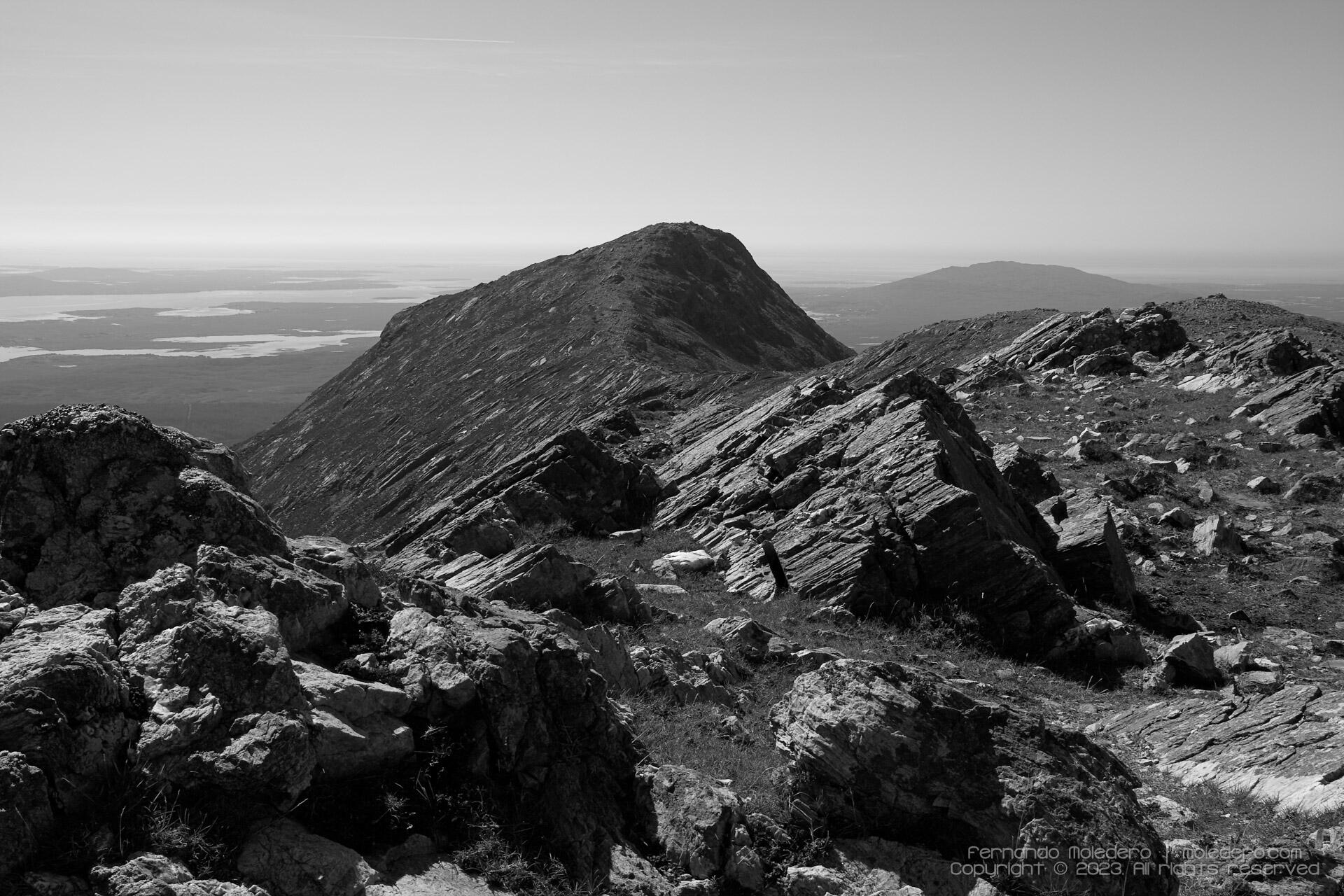 Black and white photograph of rugged rocks leading to the peak of Binn Leitri in Connemara, Ireland, with lakes and distant hills on the horizon