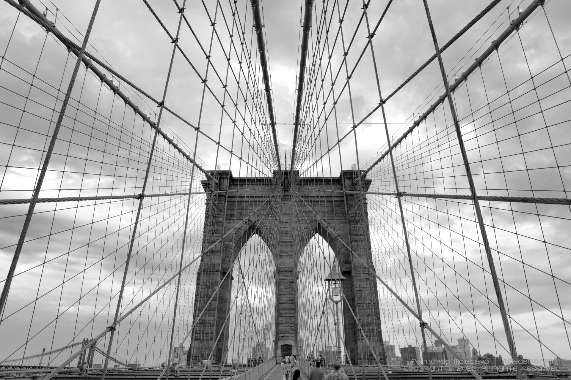 Black and white architectural composition of the Brooklyn Bridge in New York, USA, showing the stone arches and steel cables forming a symmetrical geometric pattern
