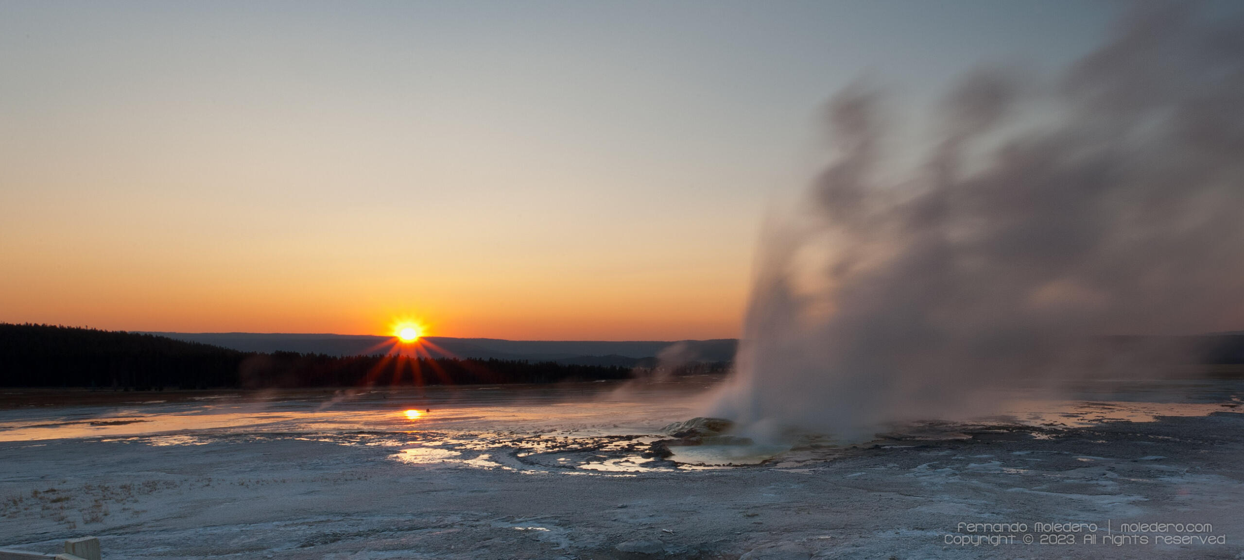 Sunrise view of the Midway Geyser Basin in Yellowstone National Park, USA, with a geyser erupting and steam rising into the orange and blue morning sky