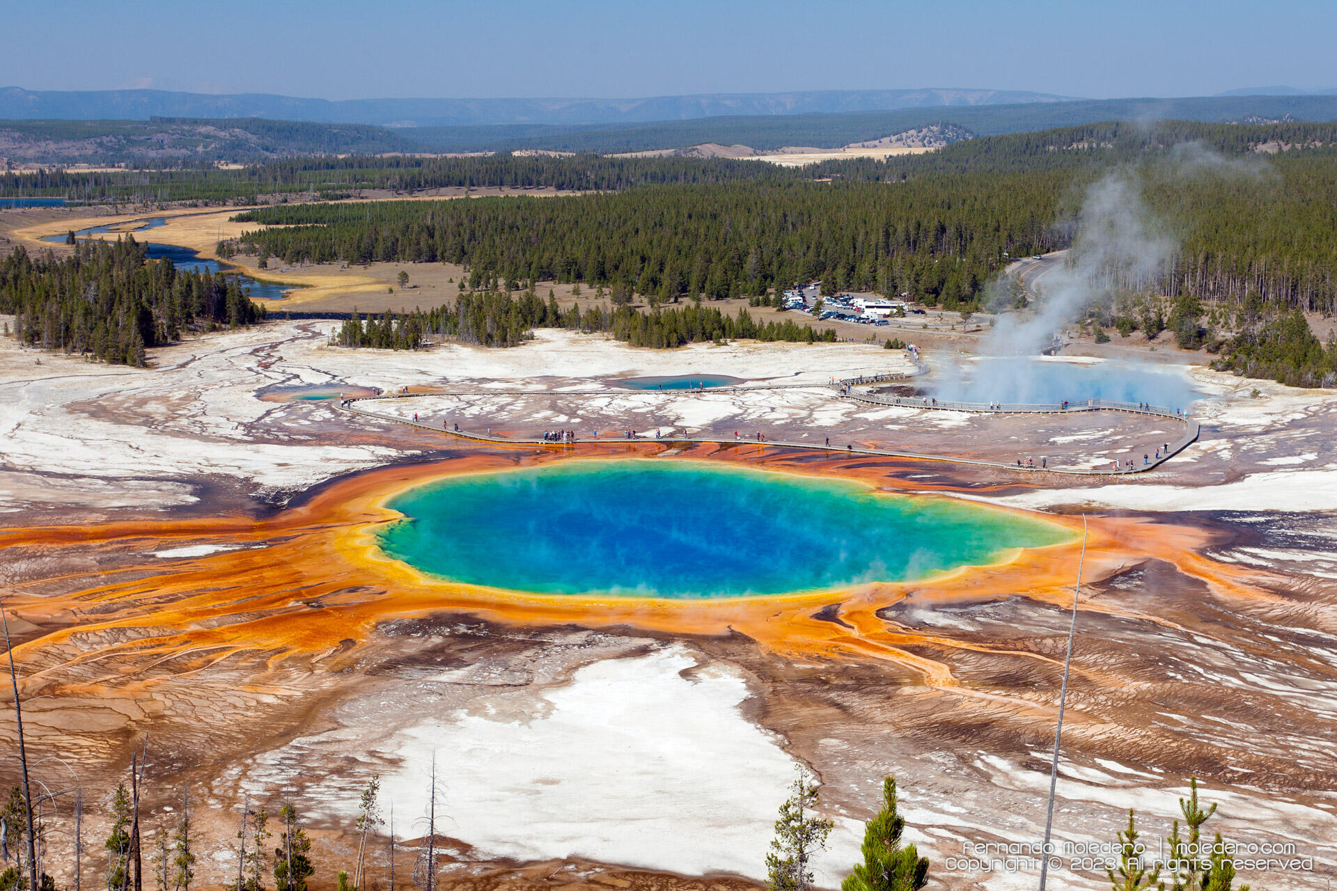 Aerial view of the Grand Prismatic Spring in Yellowstone National Park, USA, showing its vivid blue, green, yellow and orange colors surrounded by white mineral deposits, boardwalks and forest landscape