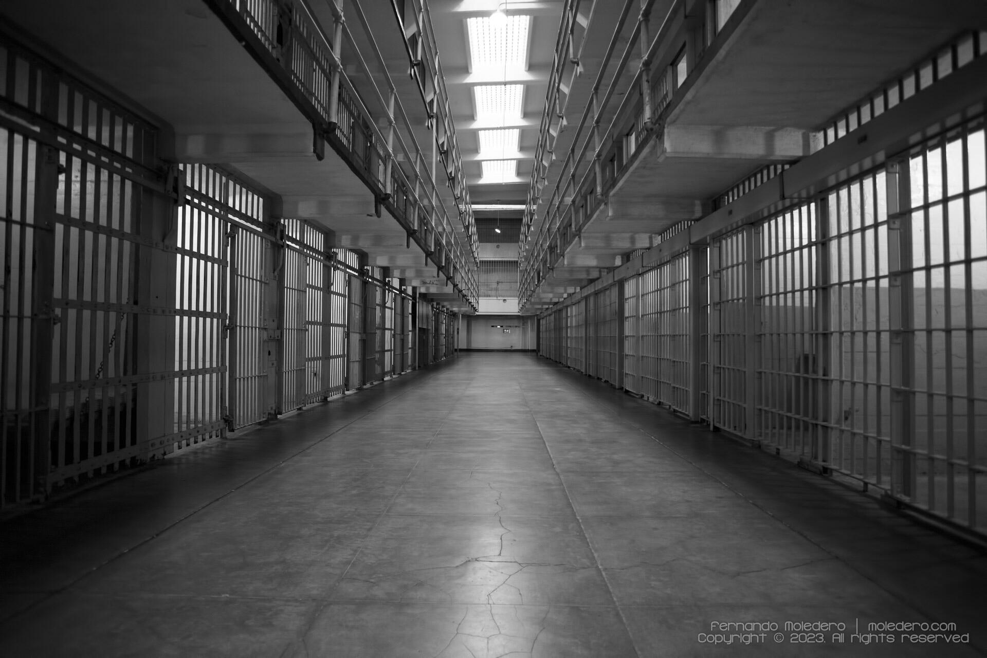 Black and white view of prison cell blocks inside Alcatraz prison in San Francisco, USA, showing symmetrical rows of barred cells and a central corridor under fluorescent lights