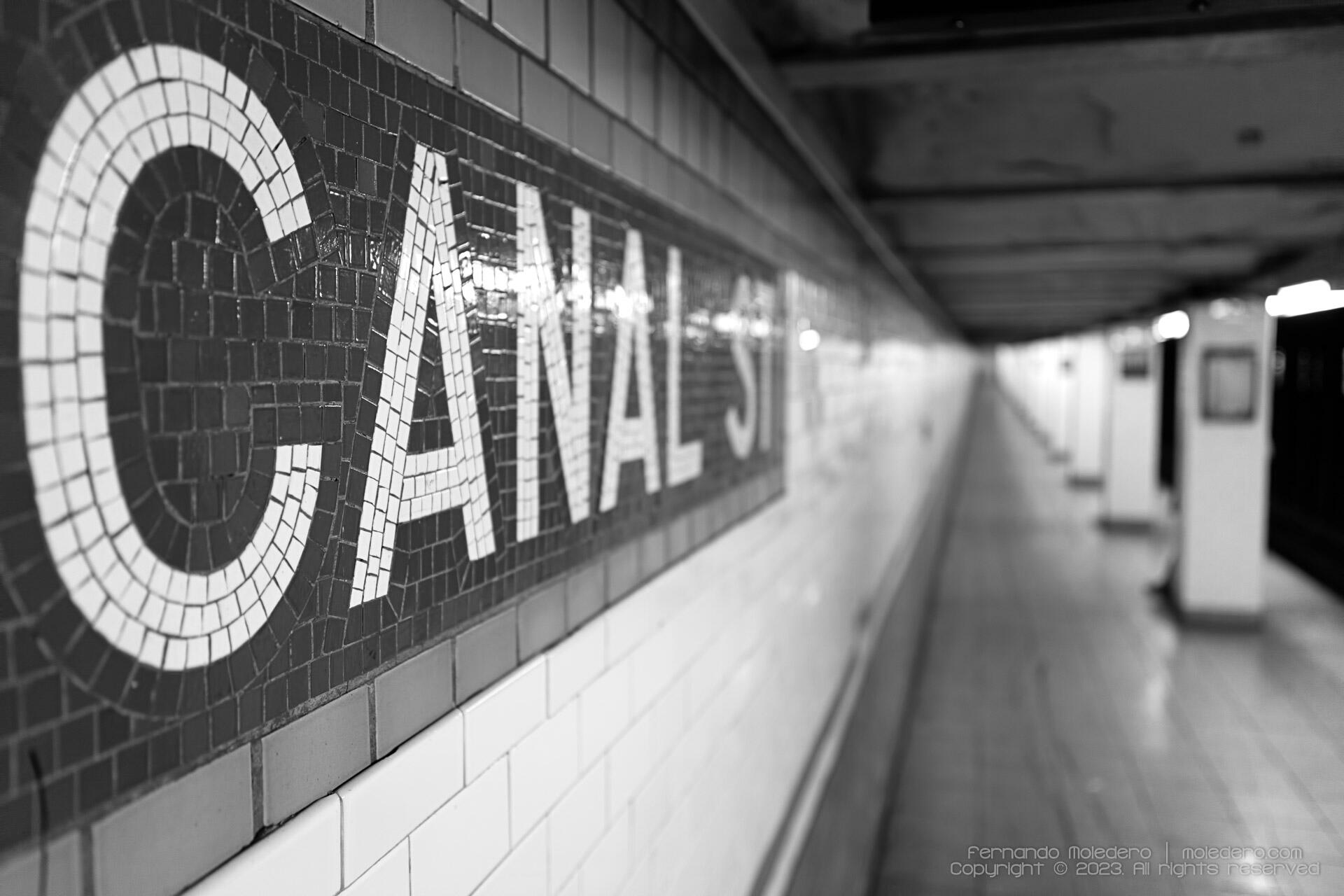 Black and white composition of Canal Street subway station in New York, USA, showing the mosaic tile sign in sharp focus with the long underground platform fading into the distance