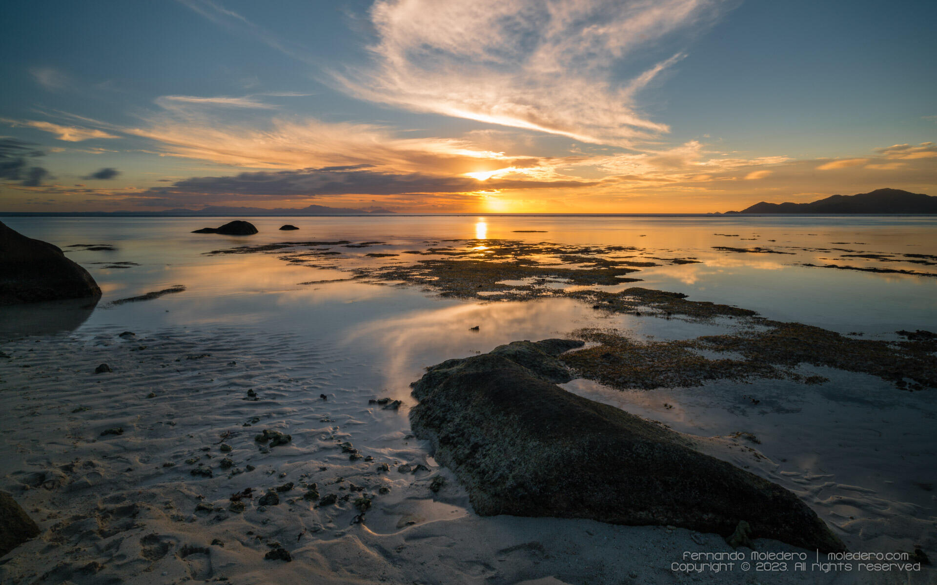 Golden sunset over Anse Source d'Argent beach in La Digue, Seychelles, with reflections on the ocean, granite rocks in the foreground and silhouettes of distant islands on the horizon