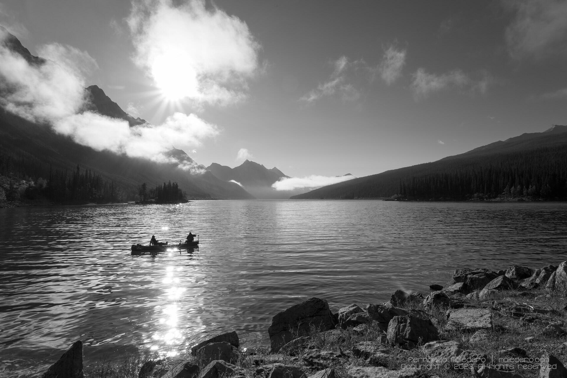 Black and white photograph of Maligne Lake in Jasper National Park, Alberta, Canada, with two people canoeing on calm waters, surrounded by forested mountains, morning mist and dramatic clouds