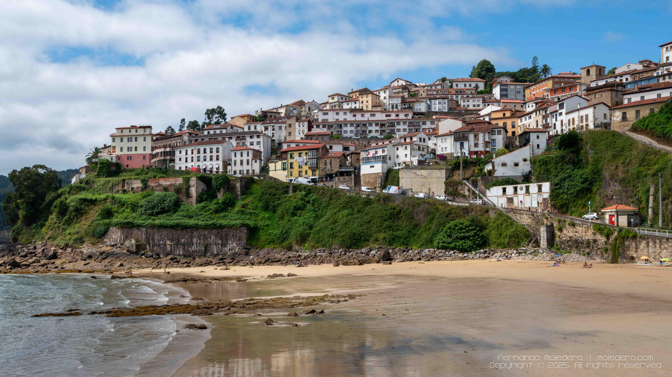 Panoramic photograph of Lastres in Asturias, Spain, showing white houses with red roofs built on a cliff above the Cantabrian Sea and the coastal beach below
