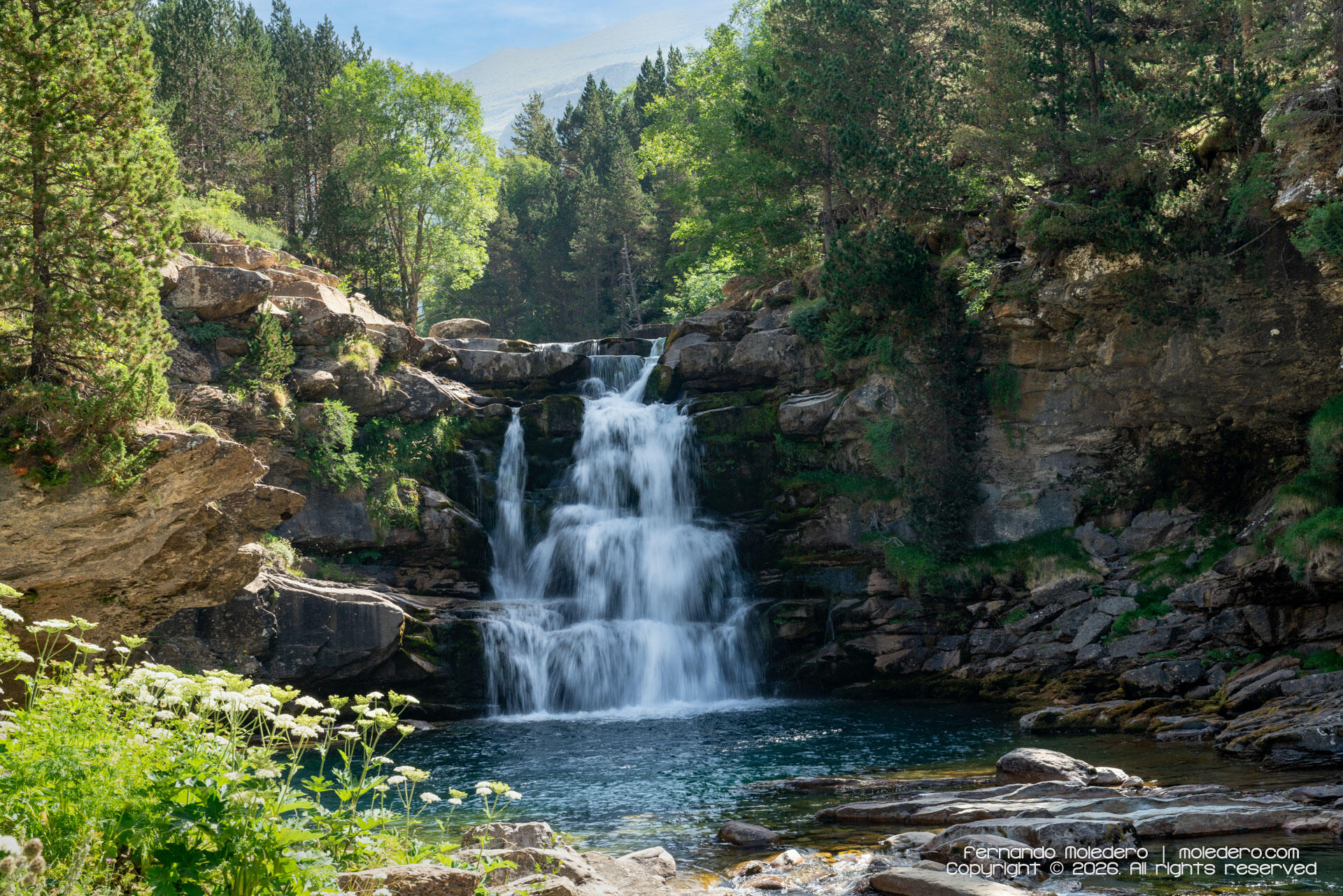 Long-exposure photograph of the Gradas de Soaso waterfalls in Ordesa Valley, Huesca, Spain, with water cascading over tiered rock formations into a turquoise mountain pool surrounded by dense forest in the Spanish Pyrenees
