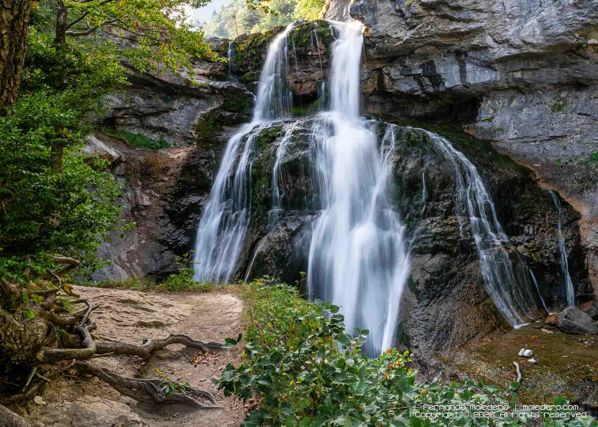 Long-exposure photograph of Cascada de la Cueva in Ordesa Valley, Huesca, Spain, with water cascading over layered limestone rocks surrounded by dense green forest in the Spanish Pyrenees.
