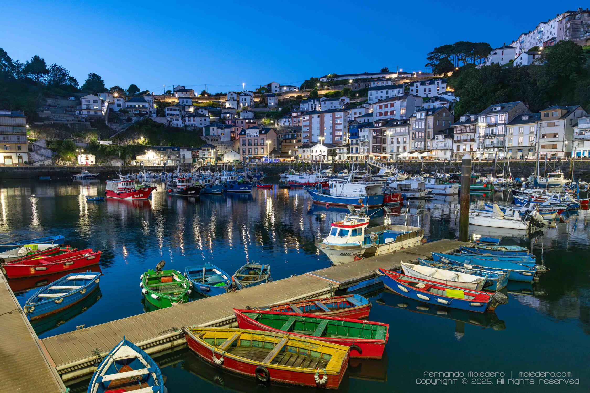 Blue hour photograph of Luarca harbor in Asturias, Spain, showing brightly painted fishing boats, illuminated hillside houses, and warm lights reflecting in the sea